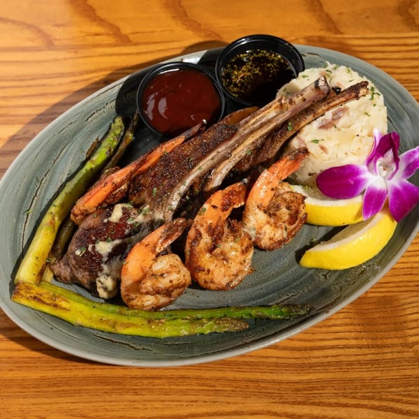 Grilled lamb chops and shrimp with mashed potatoes, asparagus, lemon, and sauces on a gray plate.