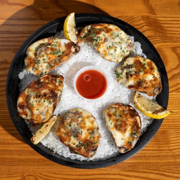 Six baked oysters with melted cheese and herbs, lemon wedges, and cocktail sauce on a bed of ice.