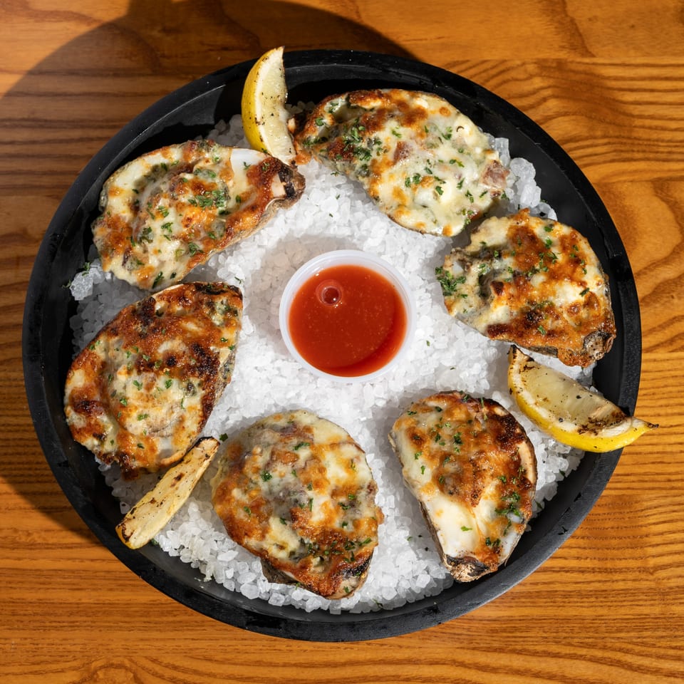 Six baked oysters with melted cheese and herbs, lemon wedges, and cocktail sauce on a bed of ice.