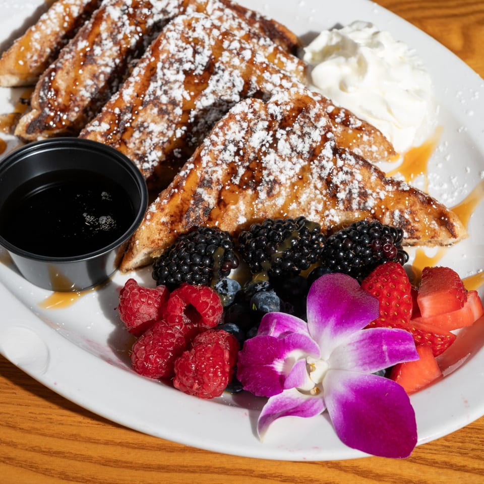 French toast with powdered sugar, syrup, whipped cream, berries, and an orchid on a white plate.